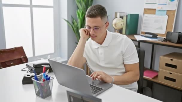 Een gefocuste jonge Spaanse man werkt op een laptop in een moderne kantooromgeving, met de nadruk op productiviteit en professionaliteit. - Video