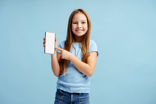Ritratto di bambina felice che indossa abiti casual con in mano il cellulare e indica uno schermo vuoto isolato su sfondo blu. Concetto tecnologico  - Foto, immagini