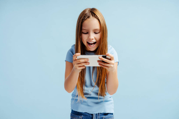 Ritratto di bambina felice che indossa abiti casual con in mano il cellulare, gioco in piedi isolato su sfondo blu. Concetto tecnologico  - Foto, immagini