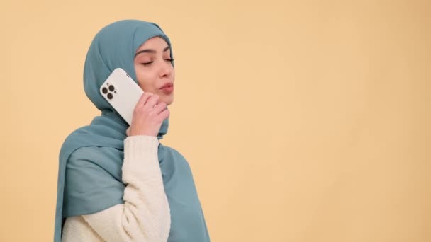 Iloinen arabinainen osallistuu keskusteluun puhelimessa lämmintä beigeä taustaa vasten, säteilee onnea ja luo ilahduttavan hetken, joka on vangittu ihastuttavassa ympäristössä.. - Materiaali, video