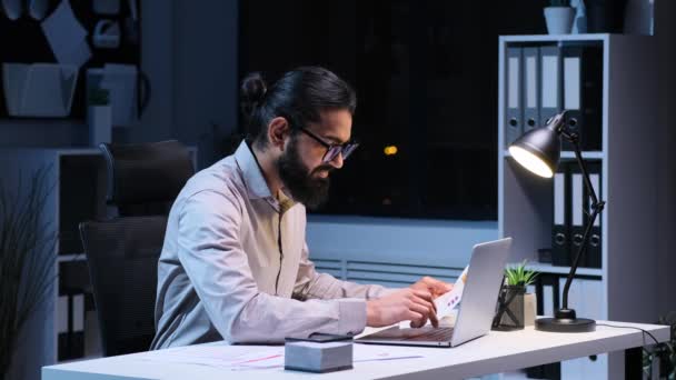 Індійський бізнесмен старанно працює з паперами та ноутбуком. Професійно одягнені, зосереджені вирази передають відчуття ефективності та відданості. - Кадри, відео