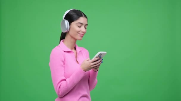 Mujer caucásica, adornada con auriculares, se captura mensajes de texto en el teléfono contra un telón de fondo verde vibrante. Su expresión enfocada y comportamiento multitarea transmiten un sentido de conectividad moderna. - Imágenes, Vídeo