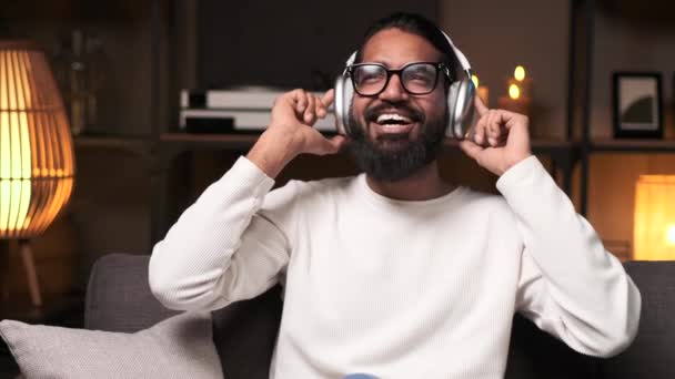 Wesoły Indianin lubi słuchać muzyki przez słuchawki w salonie. Jego uśmiechnięte miny tworzą pogodną scenę osobistej przyjemności w komfortowej atmosferze nocy.. - Materiał filmowy, wideo
