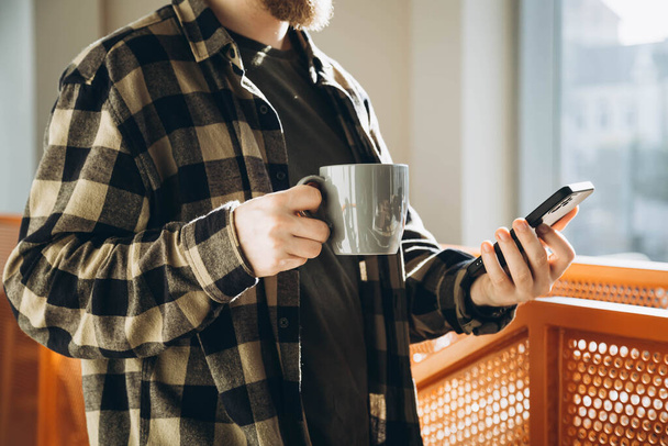 Hombre con una taza de café y teléfono inteligente por la mañana en una oficina moderna. Un joven usa un smartphone y bebe café en una cafetería. - Foto, imagen