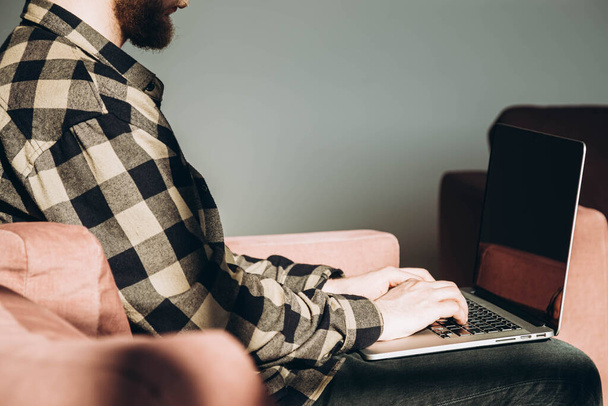 Um especialista em TI masculino trabalha remotamente, sentado com um laptop em um sofá em um quarto ensolarado. Um jovem freelancer masculino trabalha online. Estude online. Estudante com laptop.Passive lifestyle. - Foto, Imagem