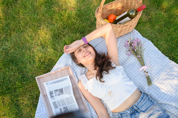 Güzel genç Asyalı kadın güneşli yaz gününde çiçek, sepet, meyve ve gazeteyle piknik örtüsünde uzanıyor. - Fotoğraf, Görsel