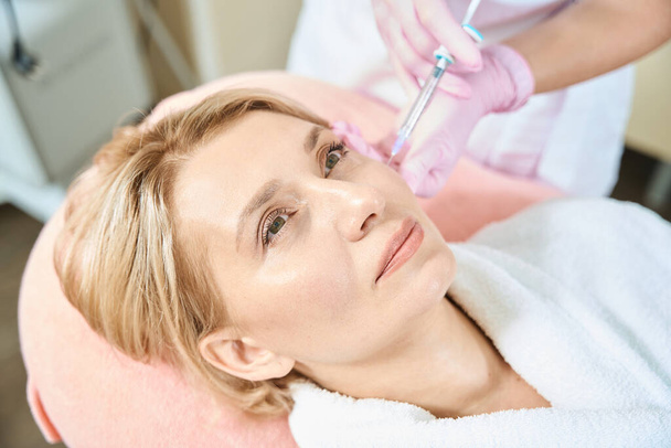 Partial female beautician doing face beauty injection with syringe of adult caucasian woman in blurred beauty salon. Face skin care, lifting and rejuvenation - Photo, Image