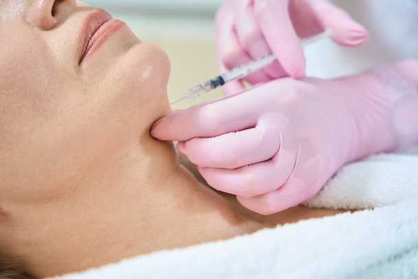 Cropped image and close up of female beautician doing face beauty injection with syringe of obscure face adult woman in blurred beauty salon. Face skin care, lifting and rejuvenation - Photo, Image