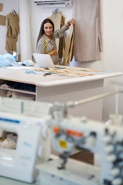Беременная ремесленница на своем рабочем месте в швейной мастерской, она использует шаблоны и ноутбук в своей работе - Фото, изображение