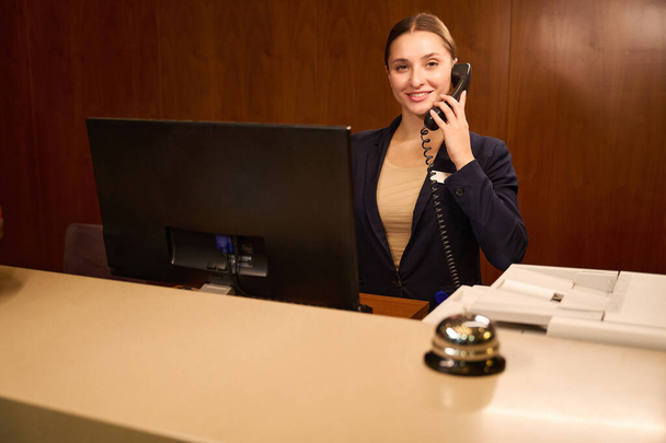 Welcoming administrator talking on phone while standing in front of computer at reception desk in hotel - Photo, Image
