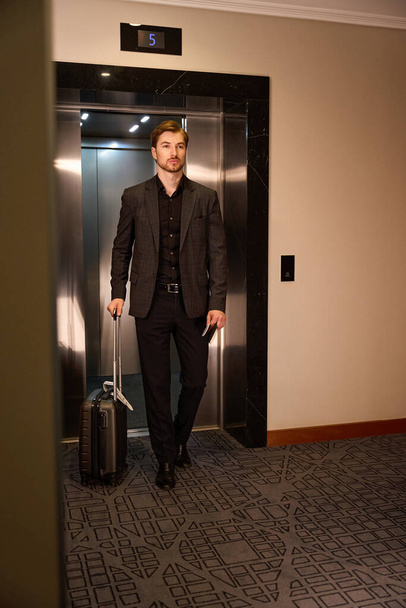 Jovem calmo saindo do elevador no hotel enquanto segura a mala em uma mão e documenta em outra - Foto, Imagem