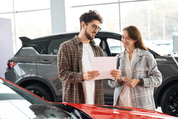 Joyful man and woman standing at dealership choosing automobile to buy reading documents - Photo, Image