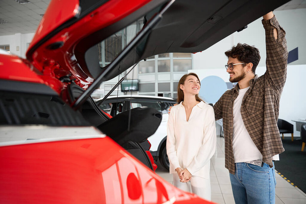 Hombre y mujer satisfechos comprando un automóvil nuevo en la concesionaria mirando el maletero de apertura del coche - Foto, Imagen