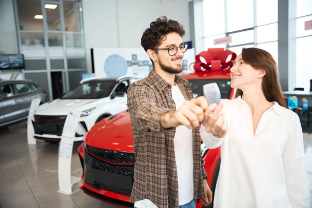 Счастливая женщина и мужчина показывают ключи от машины, празднуя покупку нового автомобиля в дилерском центре - Фото, изображение