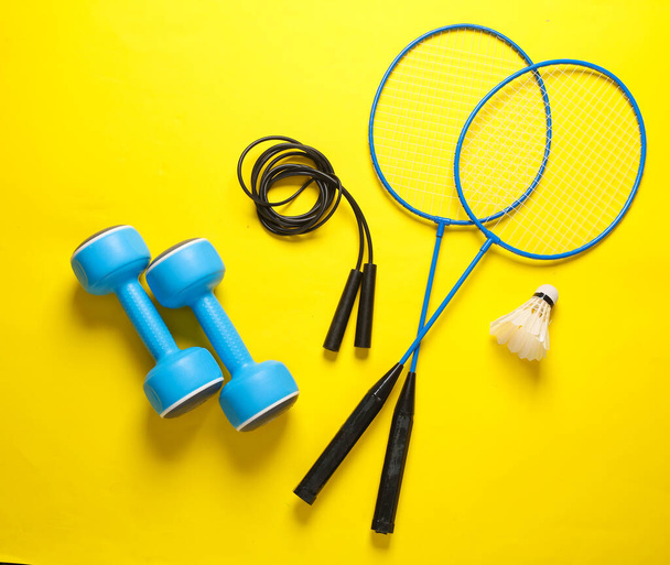 Fitness en outdoor activiteiten. Badminton racket met shuttlecock en halters springtouw op gele achtergrond. Vlakke plaat - Foto, afbeelding