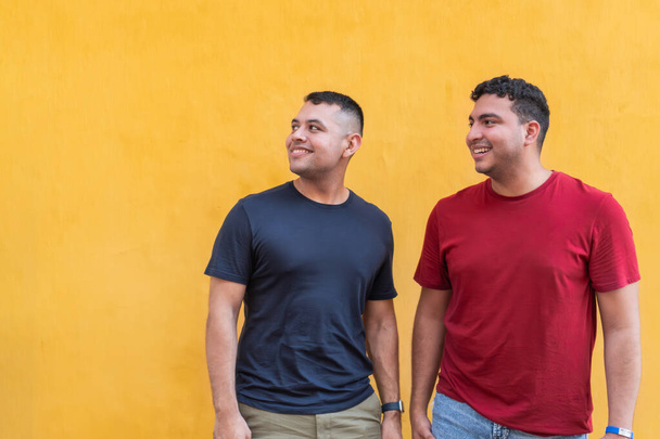 Envolvidos e felizes, dois homens de camisas coloridas olham para cima com sorrisos, em pé diante de um impressionante pano de fundo amarelo. - Foto, Imagem