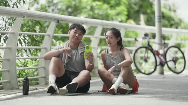 felice giovane coppia asiatica seduta a terra all'aperto guardando le foto del cellulare insieme mentre si prende una pausa dall'esercizio - Filmati, video