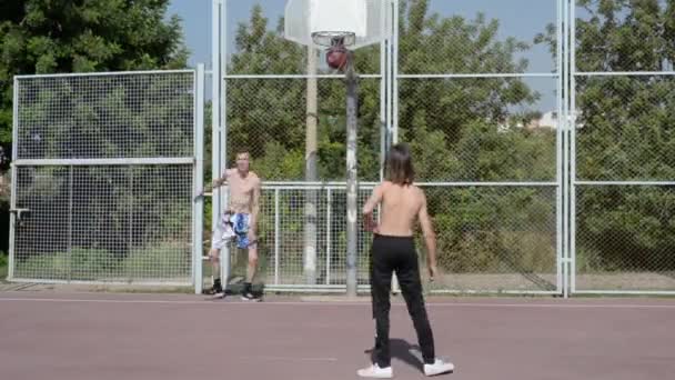 Un adolescente y un joven están jugando baloncesto en una cancha abierta en un día caluroso. Los niños lanzan una pelota en un aro. Jóvenes deportistas cerca de la casa - Imágenes, Vídeo