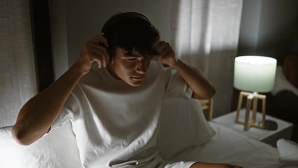 Knappe Spaanse tiener geniet van zijn favoriete lied, ontspannen in zijn gezellige slaapkamer, loungen op zijn comfortabele bed 's nachts met het schemerige licht van de lamp - Video