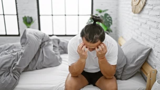 Angst grijpt jonge Chinese man, worstelend met een stressvol probleem, kijkt bezorgd, zittend op bed in een sombere slaapkamer ochtend, tekenen van emotionele nood manifesteren. - Video