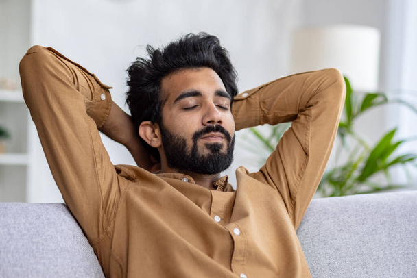 Hombre barbudo relajado con los ojos cerrados descansando en un sofá, las manos detrás de la cabeza en una habitación acogedora, concepto de paz y tiempo libre en casa. - Foto, Imagen
