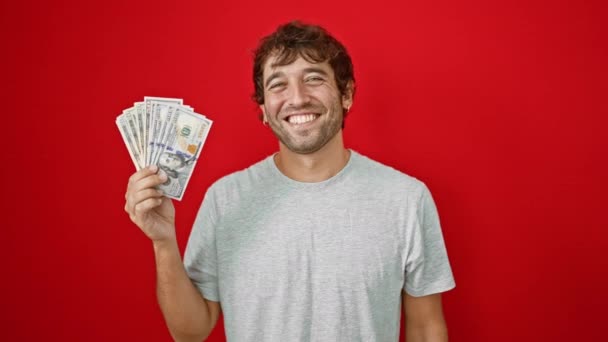 Cheerful young man, brimming with confidence, clutching american us banknotes with a joy-filled toothy smile, radiates positivity, on isolated red background. - Footage, Video