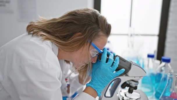 Határozott fiatal szőke tudós nő teljesen lekötött, aprólékosan dolgozik mikroszkóppal a laboratóriumban - Felvétel, videó