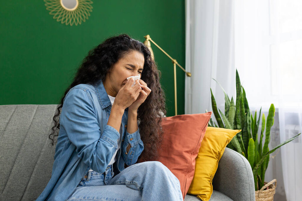 Mujer con camisa de mezclilla sintiéndose mal, estornudando en el tejido en el sofá, interior del hogar con plantas, expresando síntomas de enfermedad y alergia. - Foto, imagen