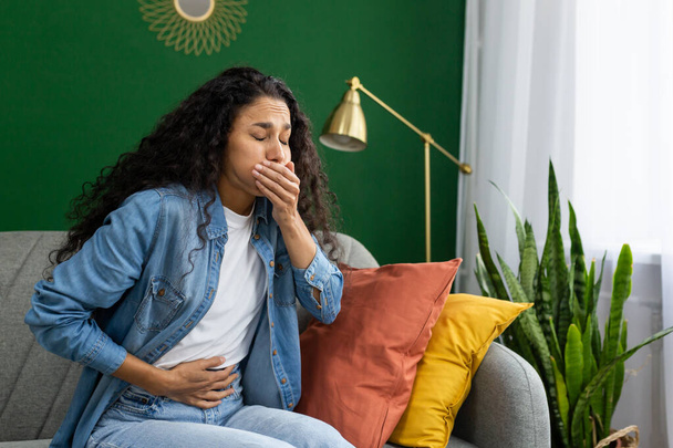 Una giovane donna afflitta sensazione di nausea su un divano, mano sulla bocca, cercando di sopprimere la voglia di vomitare. - Foto, immagini