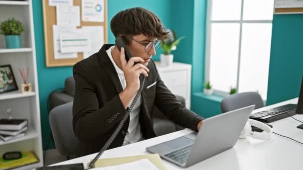 Joven adolescente hispano trabajador de negocios hablando por teléfono usando portátil quitándose las gafas en la oficina - Imágenes, Vídeo