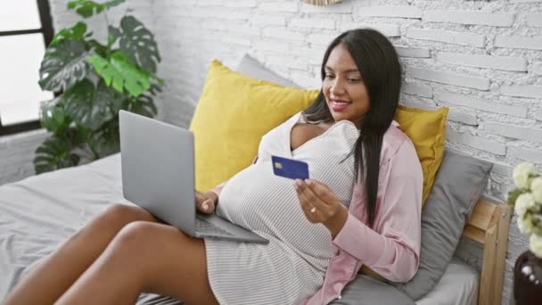 Sonriendo joven madre embarazada disfrutando de su mañana, descansando cómodamente en la cama en el dormitorio, entregándose a las compras en línea con tarjeta de crédito en su computadora portátil - Metraje, vídeo