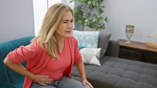 Een vrouw van middelbare leeftijd ervaart buikpijn terwijl ze in haar moderne woonkamer zit en een gezondheidsthema uitdraagt. - Video
