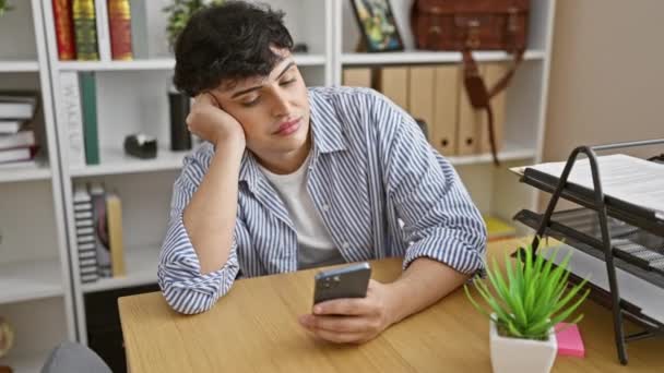 Un joven con una camisa a rayas parece aburrido con un teléfono inteligente en una oficina moderna. - Imágenes, Vídeo