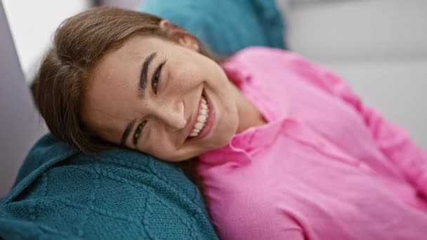 Säteilevä nuori latino nainen osoittaa iloa, luottamusta, rentoutumista istuen mukavasti hymyillen mukavalla sohvalla kauniissa kodissaan sisätiloissa. - Materiaali, video