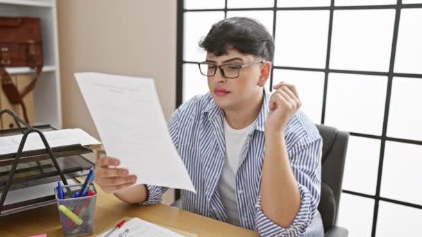 Νεαρός με γυαλιά διαβάζει έγγραφα και δείχνει απογοήτευση σε ένα σύγχρονο περιβάλλον γραφείου. - Πλάνα, βίντεο
