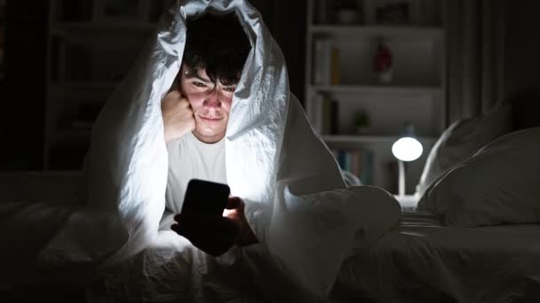 Knappe jonge Spaanse tiener die terloops in bed ligt, verscholen onder een knusse deken in de donkere slaapkamer, geabsorbeerd in een fascinerende online zoektocht op zijn smartphone, de enige lichtbron in de kamer - Video