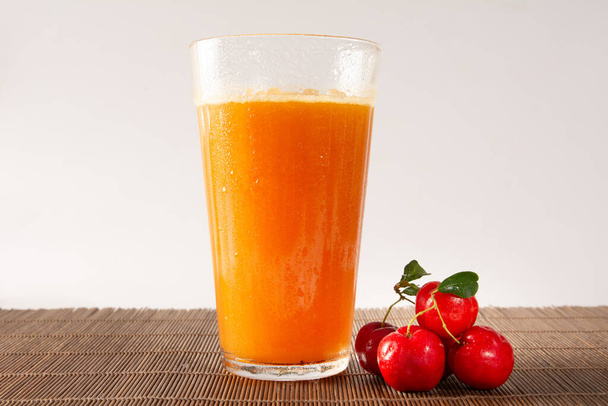 Φρέσκο βιολογικό Acerola και χυμός πορτοκαλιού σε γυάλινο κύπελλο με μούρα acerola σε λευκό καθαρό στούντιο φωτογραφία μπροστά - Φωτογραφία, εικόνα