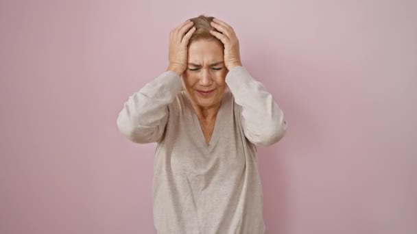 Stresszes középkorú szőke nő áll rózsaszín elszigetelt háttér, szorítja a fejét a fájdalom, sújtja a migrén fejfájás és szenvedés csendes kétségbeesés - Felvétel, videó