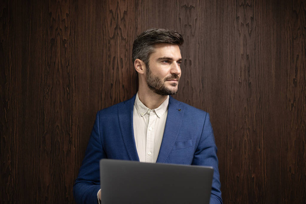 Retrato de hombre de aspecto inteligente en traje de negocios sosteniendo ordenador portátil y mirando hacia los lados. - Foto, imagen