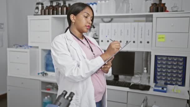 Expecting young woman scientist, deep in thought, meticulously taking notes in lab amidst medical research - Footage, Video