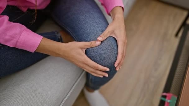 Latino naisen kädet kiinni kipeä polvi, sisätiloissa kärsivät tuskallinen vamma olohuoneen sohvalla - Materiaali, video
