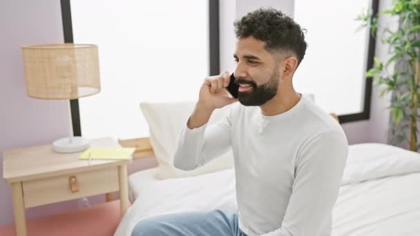 Glimlachende jonge man met een baard aan de telefoon in een moderne slaapkamer, die een casual, ontspannen levensstijl thuis portretteert. - Video