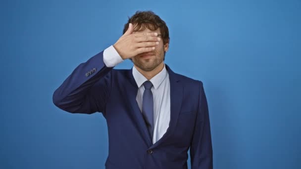 Cheerful young man in business suit, playfully covering his eyes for a blind surprise, smiling vibrantly over blue isolated background - Footage, Video