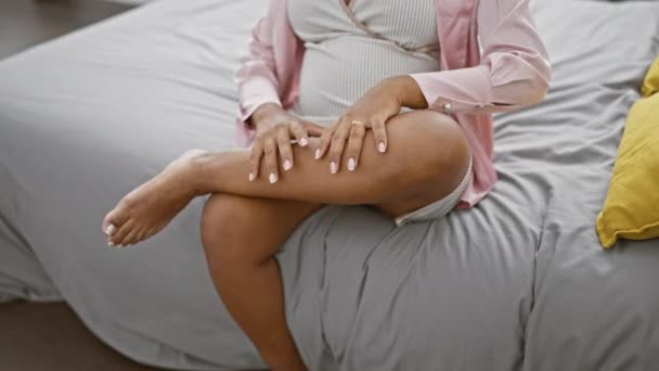 Jovem hispânica exausta esperando maternidade, sentada na cama, em casa, massageando a perna cansada em seu quarto acolhedor, tocando a barriga grávida em pijama. - Filmagem, Vídeo