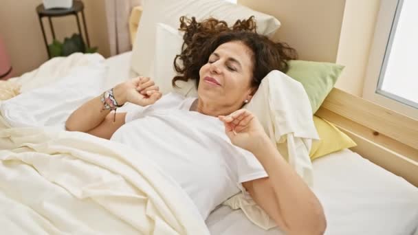 volwassen vrouw stretching in bed met witte lakens in een heldere ochtend slaapkamer instelling - Video