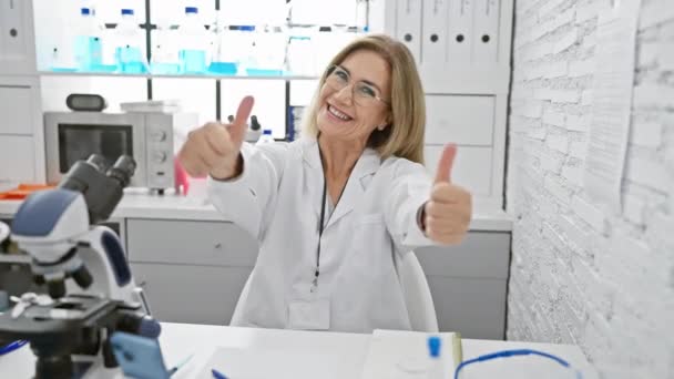 Mulher loira alegre de meia-idade cientista mostrando gesto vencedor, polegares para cima no laboratório, brilhando com felicidade, sorrindo de orelha a orelha em um momento triunfante! - Filmagem, Vídeo