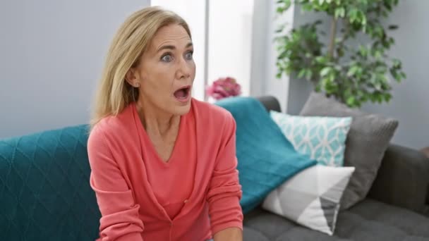 Erstaunte blonde Frau mittleren Alters, die ungläubig auf dem Sofa sitzt, den Mund vor Angst weit aufgerissen, mit einem überraschten Gesicht zu Hause in einer Inneneinrichtung - Filmmaterial, Video