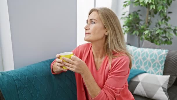 Uma mulher serena desfruta de uma caneca de chá em um sofá teal em uma sala de estar moderna. - Filmagem, Vídeo