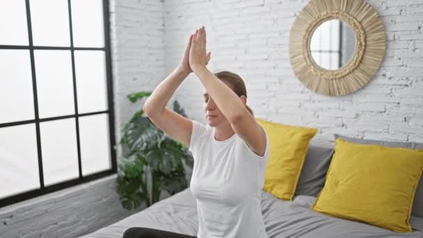Krásná, vážná mladá blondýnka ponořená do klidné rovnováhy skrze jóga cvičení na posteli, sedící v soustředěném soustředění ve svém ložnici interiéru, dopřávající si ranní relaxaci. - Záběry, video