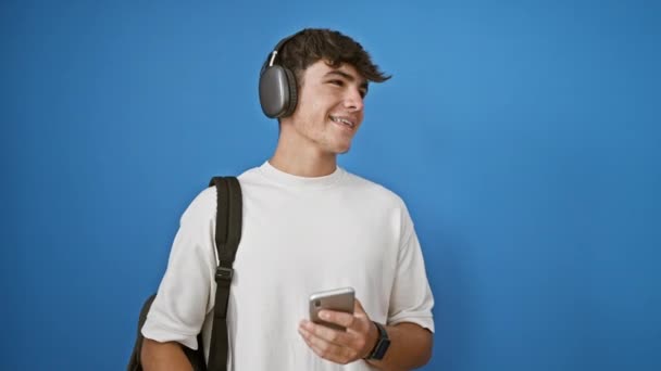 Adolescent hispanique cool dansant au rythme, étudiant occasionnel utilisant la technologie intelligente, écoutant de la musique sur un téléphone, isolé sur un fond bleu. - Séquence, vidéo